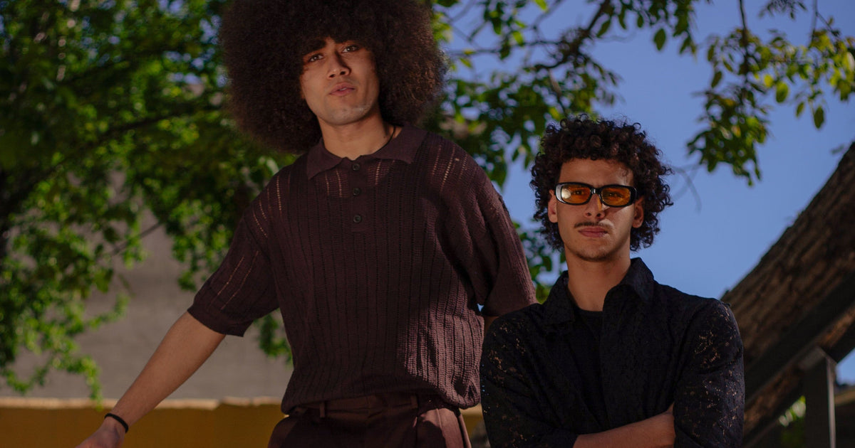 Two men standing outdoors under trees, one wearing a dark brown short-sleeve button-up shirt and brown pants, and the other wearing a black lace-sleeve shirt and orange-tinted sunglasses.
